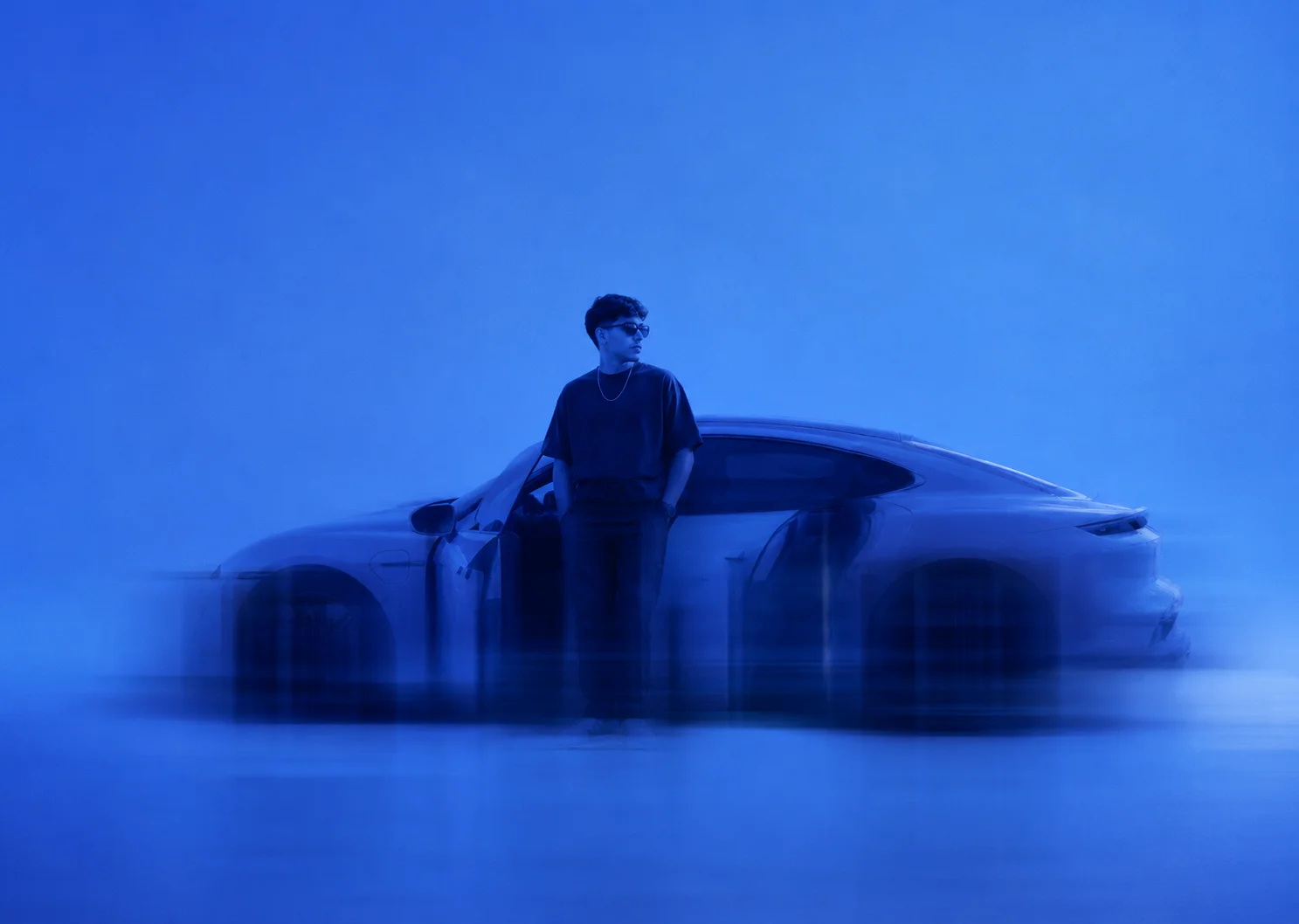 Man wearing sunglasses stands with hands in pockets beside a sleek car in a blue-tinted scene.