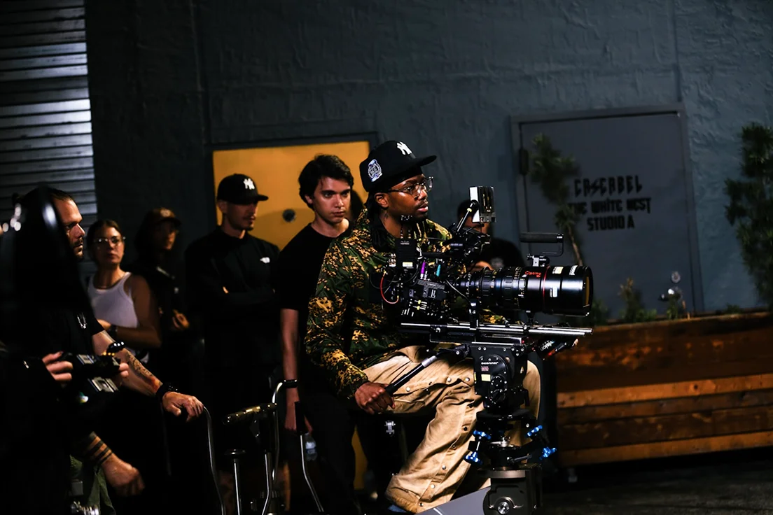 Camera operator with multiple crew members filming at night outside a studio with blue walls.