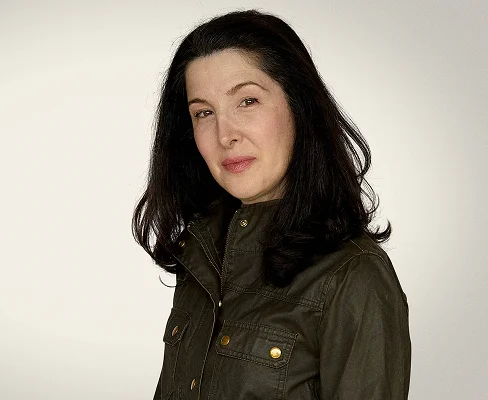 Woman with long dark hair wearing an olive jacket, looking at the camera with a slight smile.