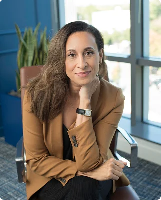 Woman with long hair and tan blazer sitting on chair in office with hand on chin, smiling gently.