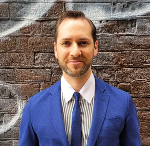 Man with short brown hair and trimmed beard wearing a blue suit and tie, standing in front of a brick wall.