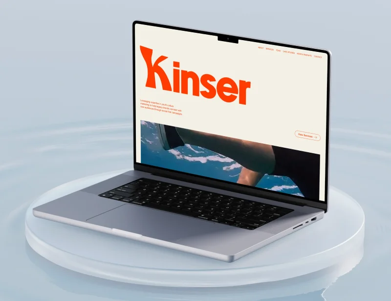 Open laptop on a round platform displaying a website with large orange Kinser text and ocean image.