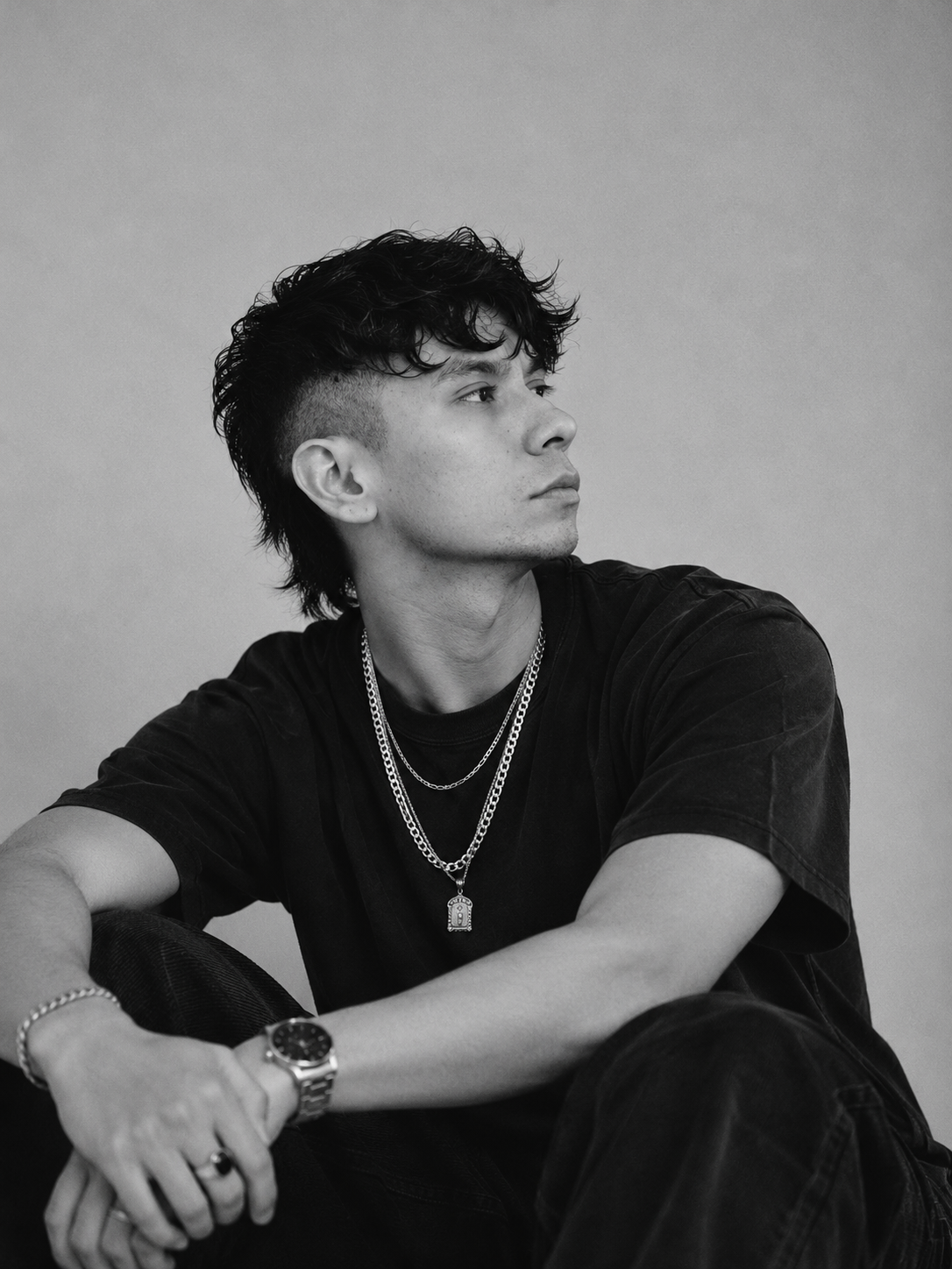 Black and white portrait of a contemplative young man with curly hair looking to the side, wearing a dark t-shirt, two chain necklaces with a pendant, a wristwatch, and a bracelet, seated with folded arms.