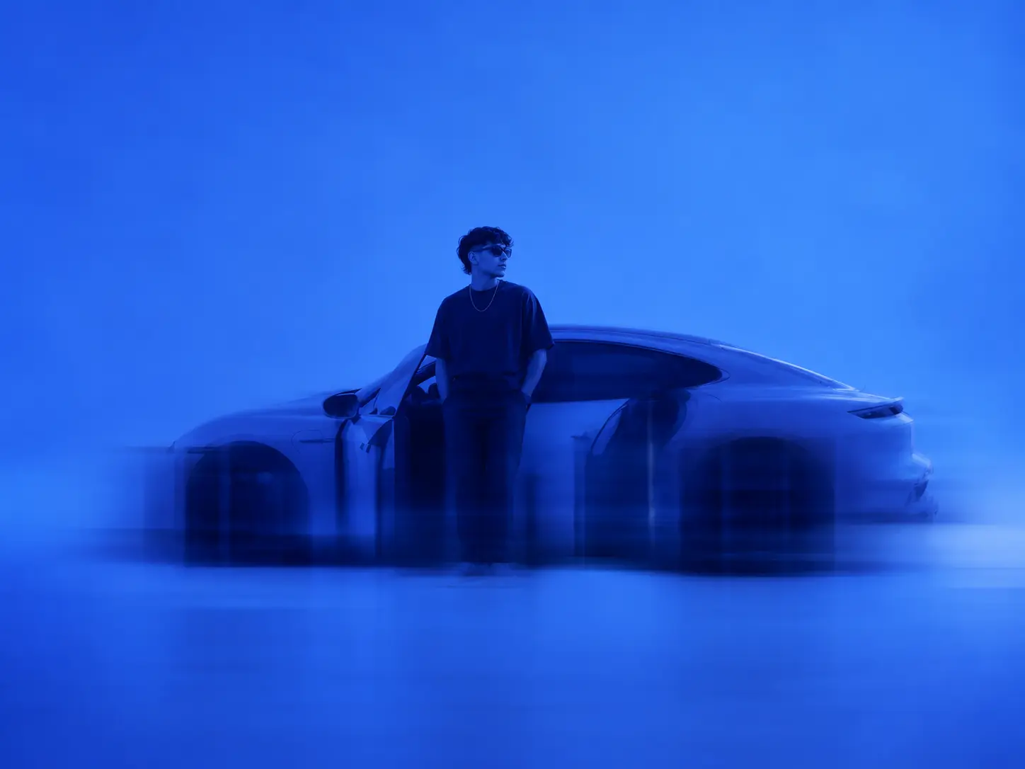 Man wearing sunglasses stands with hands in pockets beside a sleek car in a blue-tinted scene.