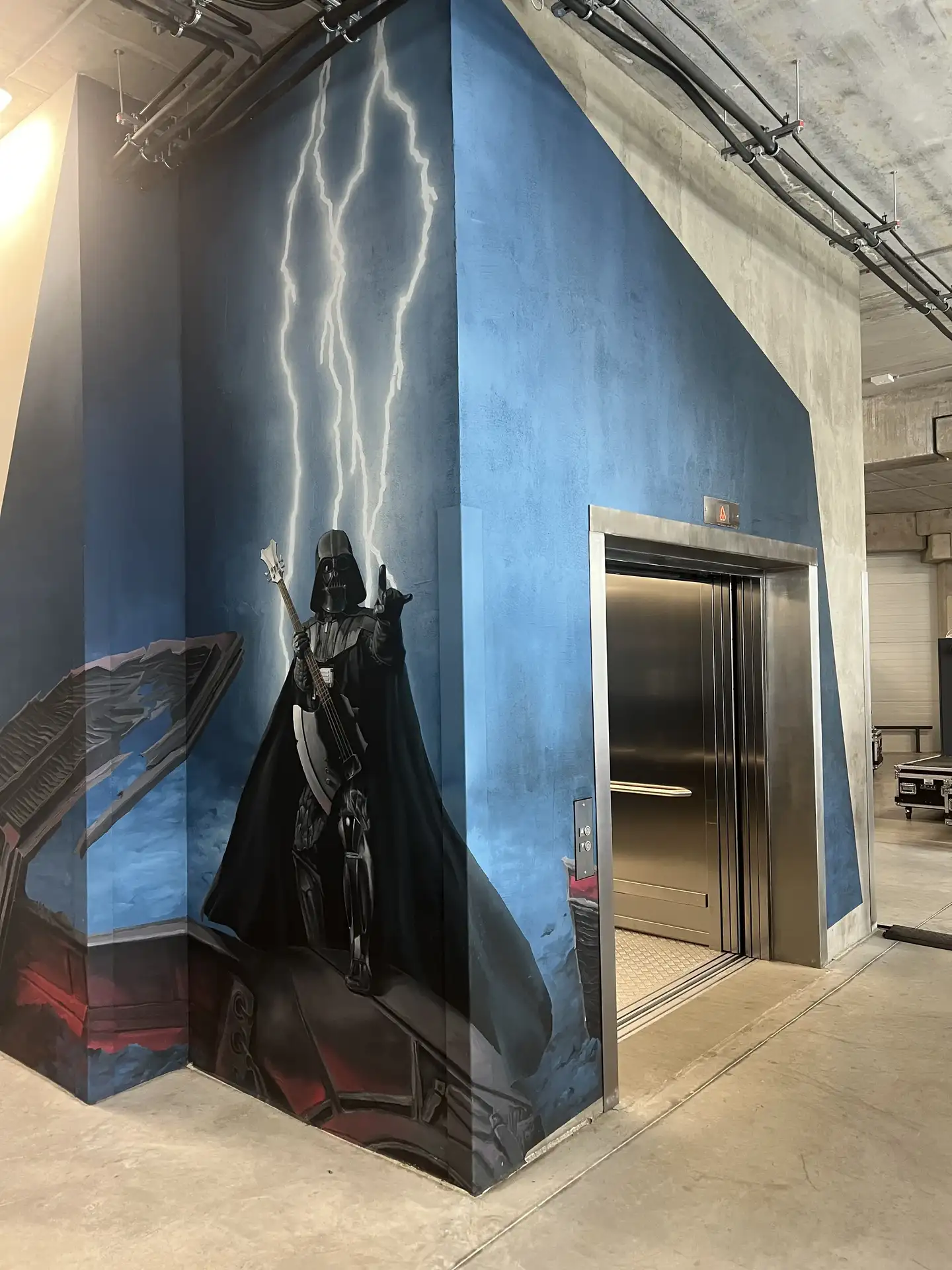 Modern ipari stílusú folyosón álló lift, amelynek falát Darth Vader villámokkal kiegészített, látványos falfestménye díszíti.