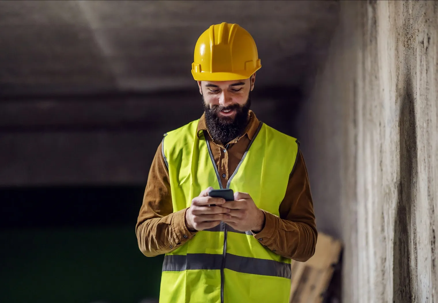 Bearded construction worker in a yellow hard hat and safety vest looking at a smartphone inside a building.