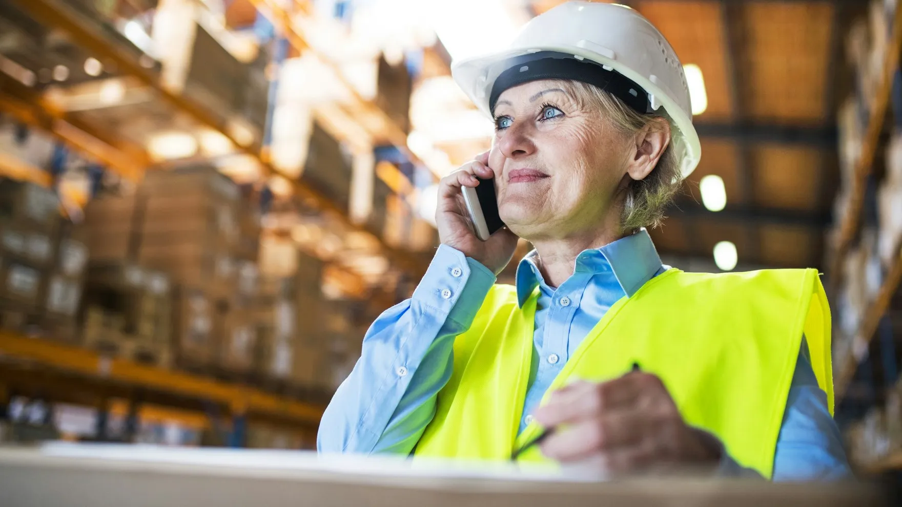 Woman wearing a white hard hat and yellow safety vest talking on a phone inside a warehouse.