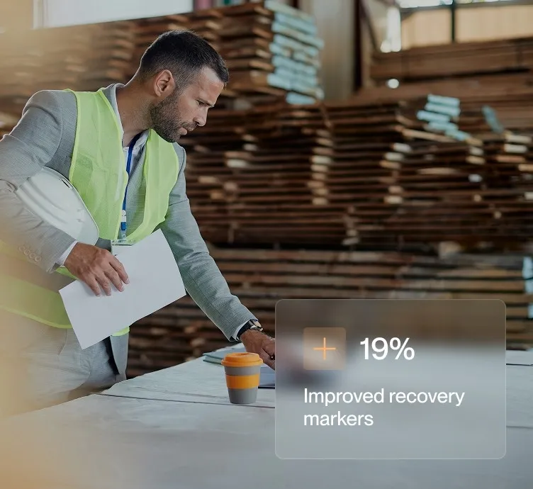 Man in safety vest and suit holding papers at a table in a wood manufacturing facility.