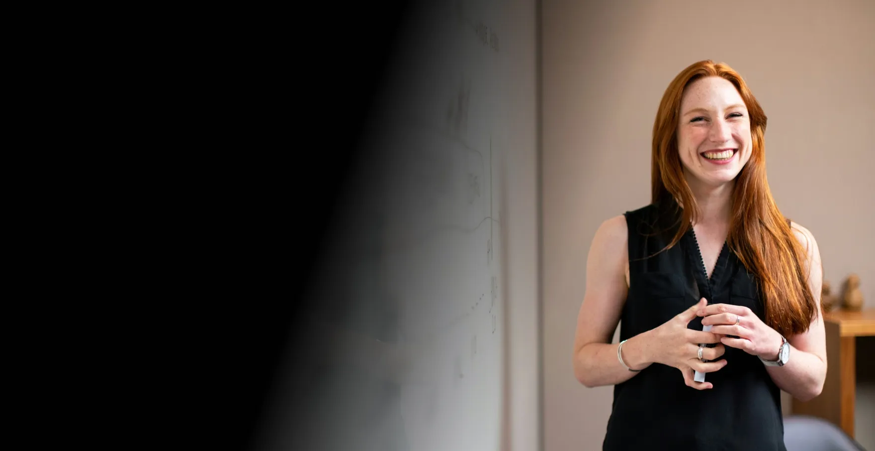 Smiling woman with long red hair stands by a whiteboard with diagrams, wearing a black sleeveless top.