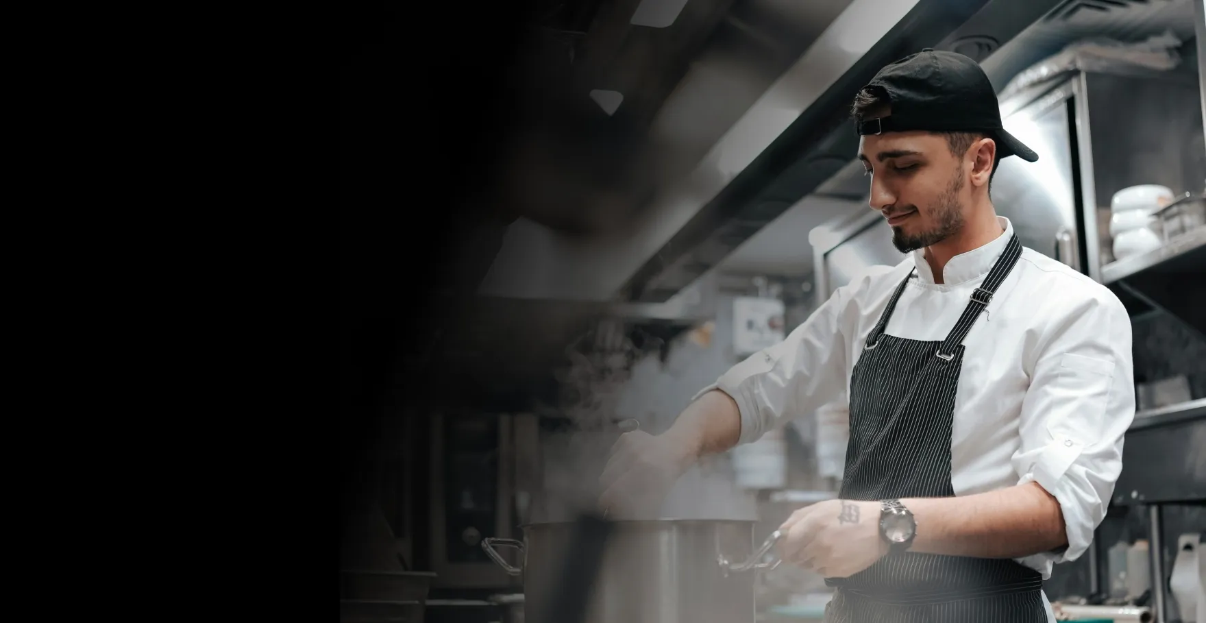 Chef in a black cap and apron stirring a steaming pot in a professional kitchen.