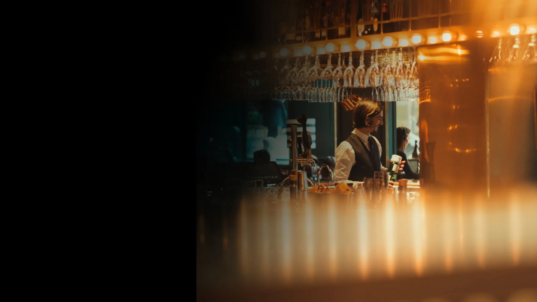 Bartender in white shirt and vest smiling while holding a bottle behind a warmly lit bar.