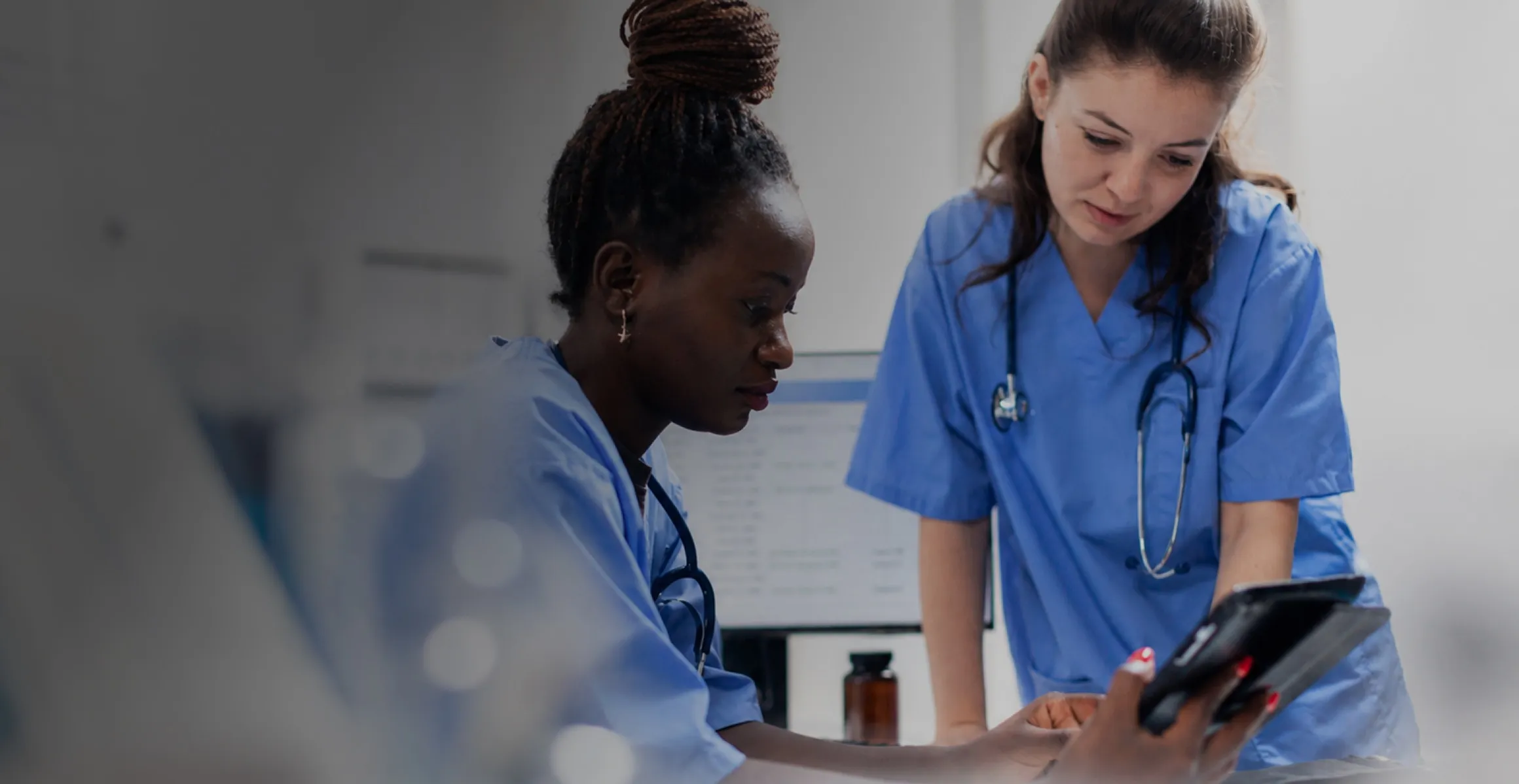 Two nurses in blue scrubs looking at a tablet together in a clinical setting.