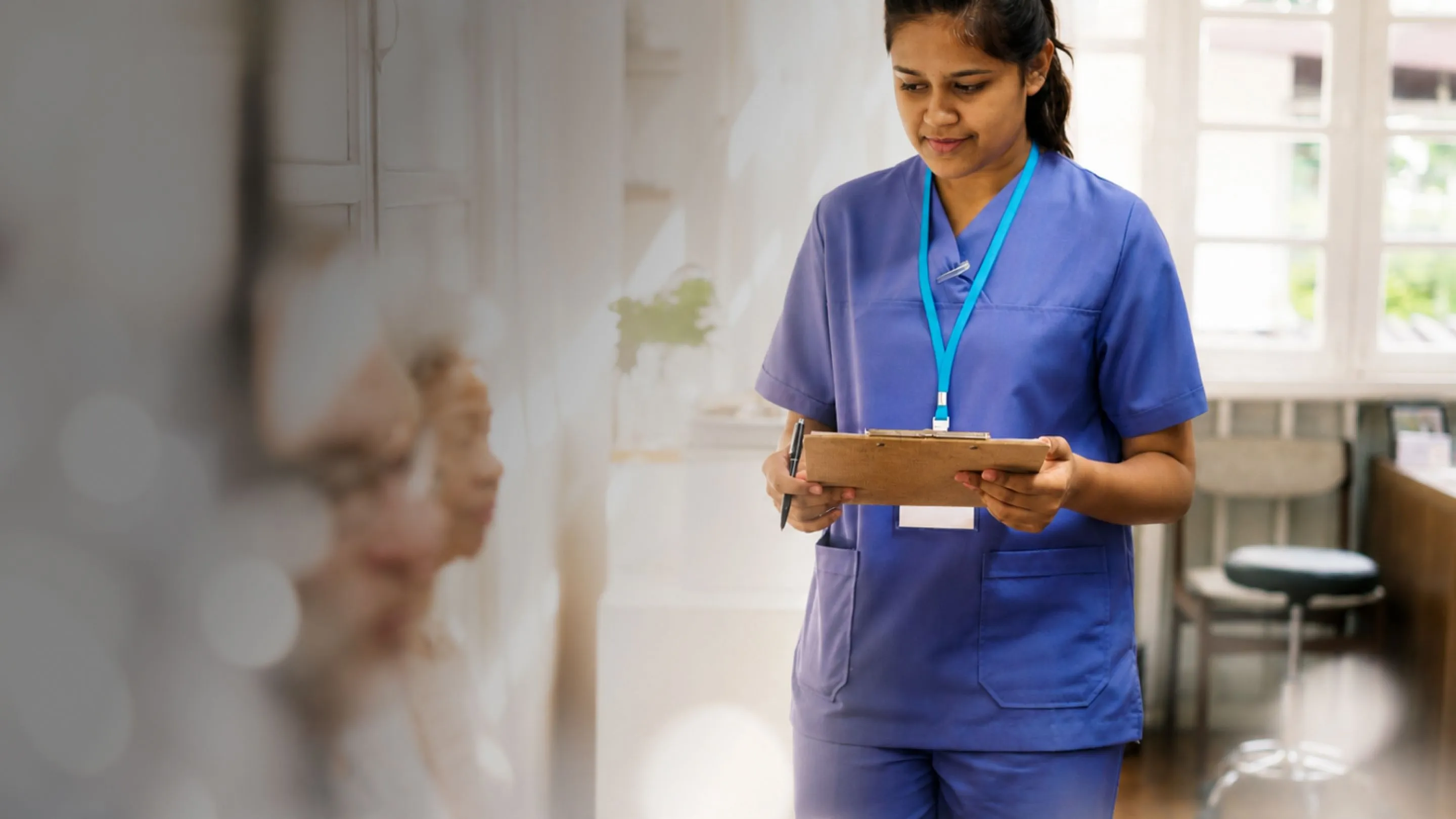 Healthcare worker in blue scrubs holding a clipboard and pen, checking on blurred elderly patients.