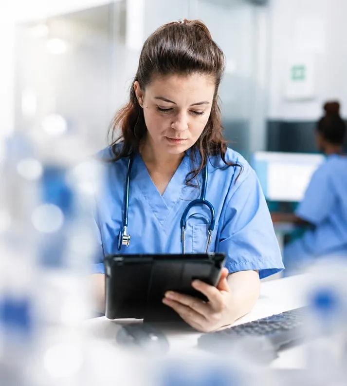 Female nurse in blue scrubs with stethoscope reviews tablet in a medical office.