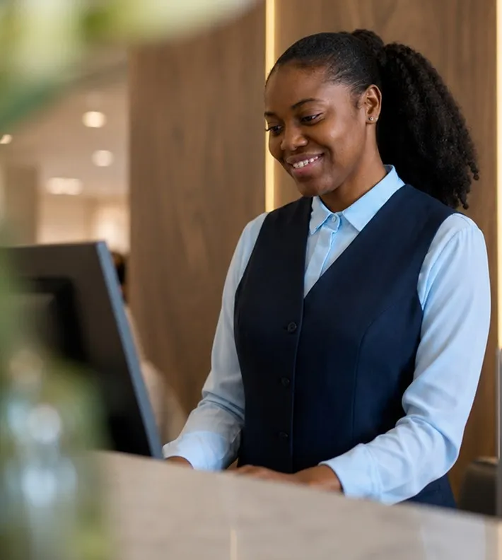 Smiling receptionist in a blue shirt and navy vest working at a computer at the front desk.