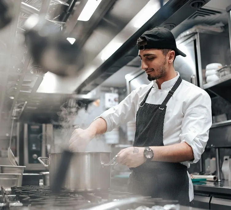 Chef wearing a black cap and apron stirring food in a steaming pot in a professional kitchen.