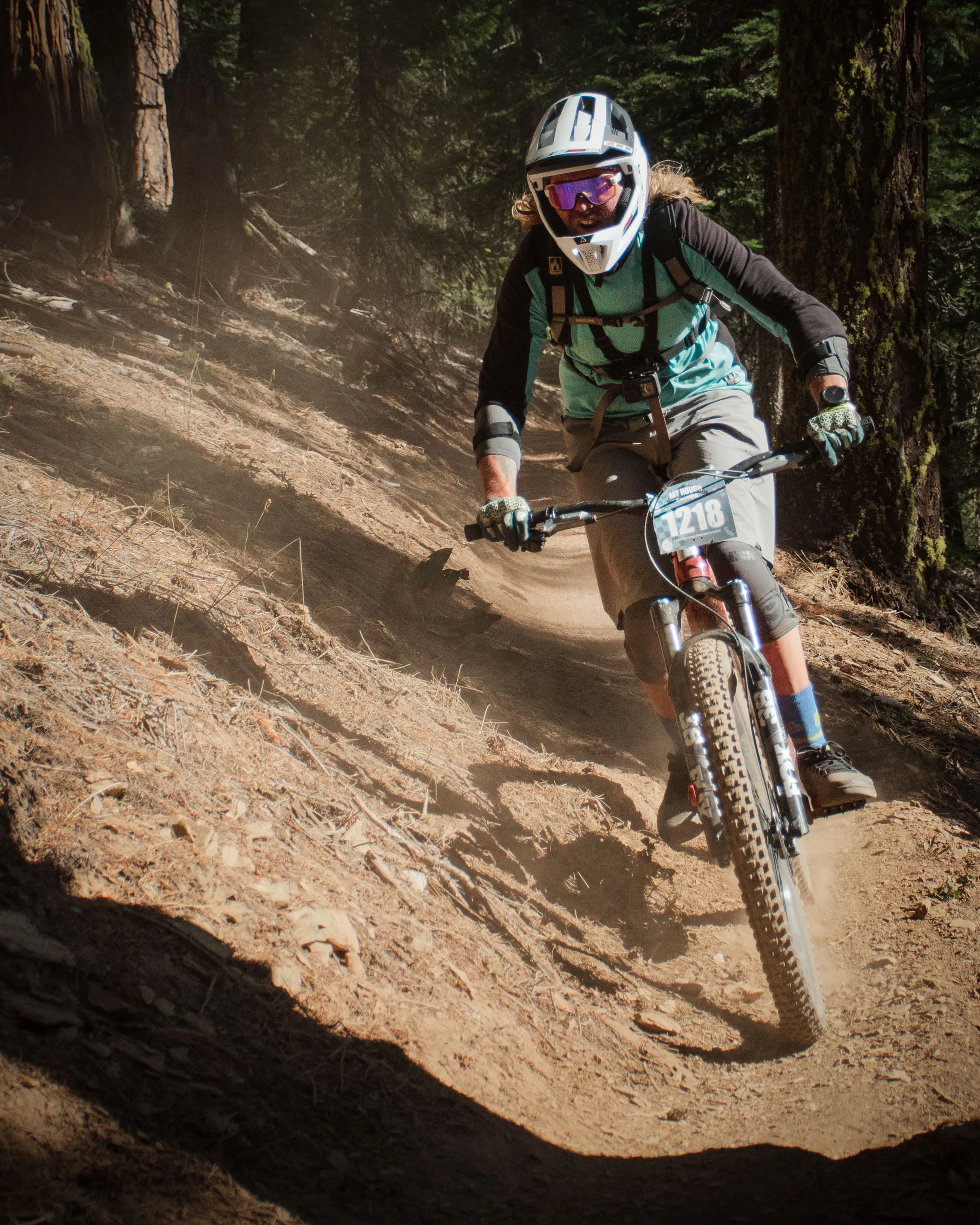 A mountain biker wearing a helmet, goggles, and protective gear rides down a dusty forest trail, leaning into a turn with dirt kicking up from the back wheel.