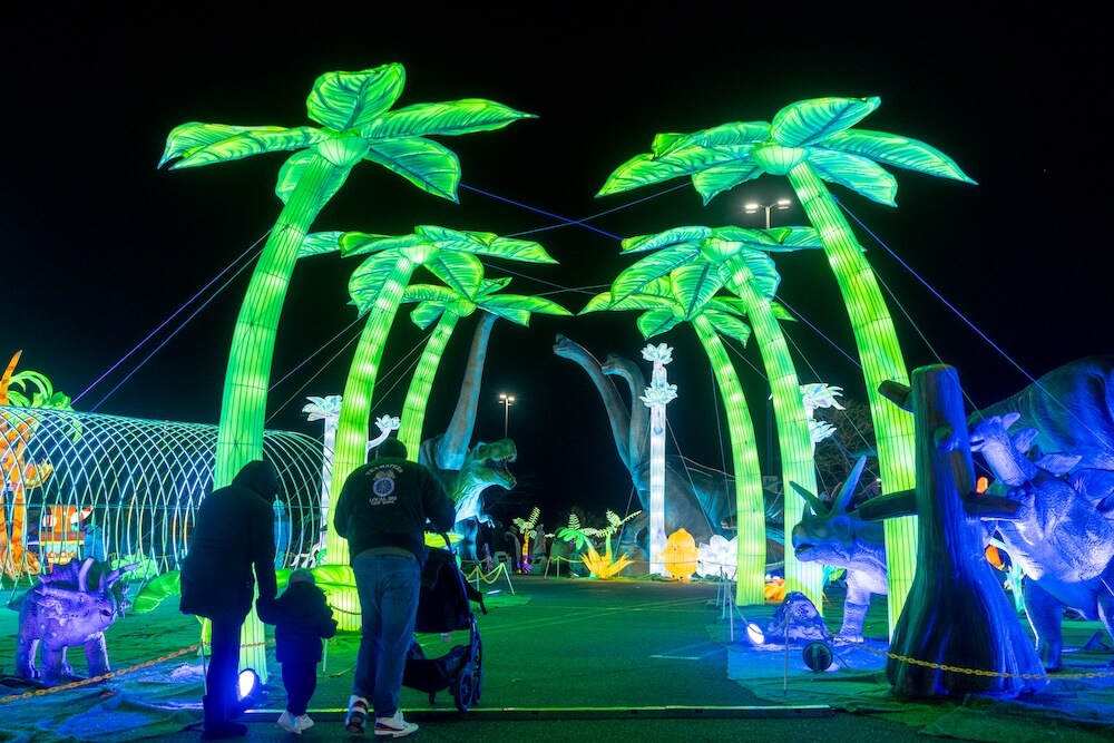 A family walking through a glowing green palm tree light tunnel surrounded by dinosaur sculptures at night during LuminoCity Dino Safari. 
