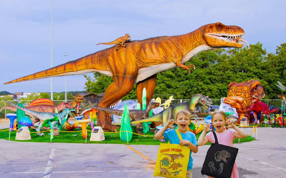 Two happy children holding dinosaur tote bags posing playfully in front of a giant T-Rex statue at LuminoCity Dino Safari.