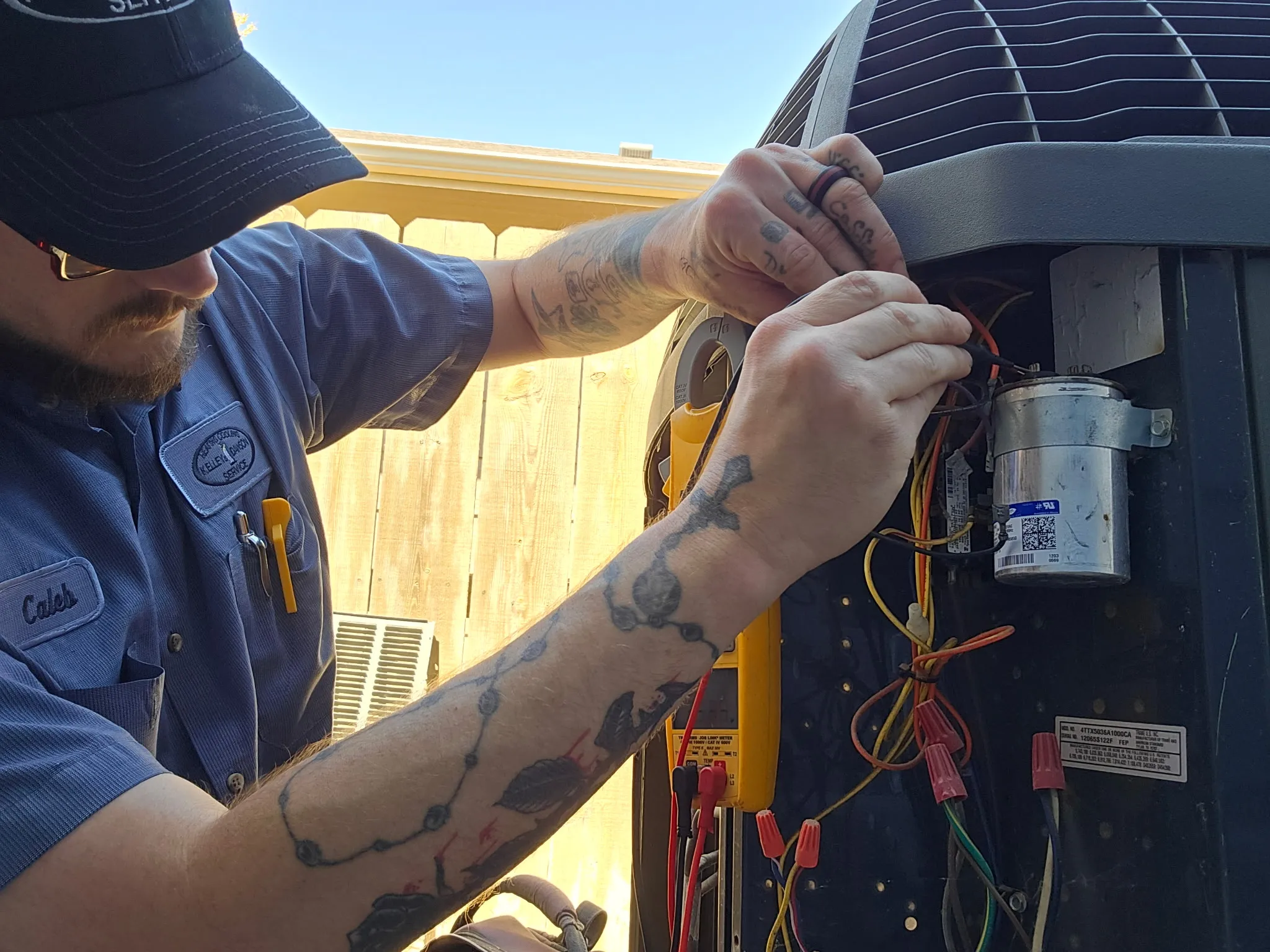 A tattooed technician in a blue uniform shirt works on an air conditioning unit outdoors.