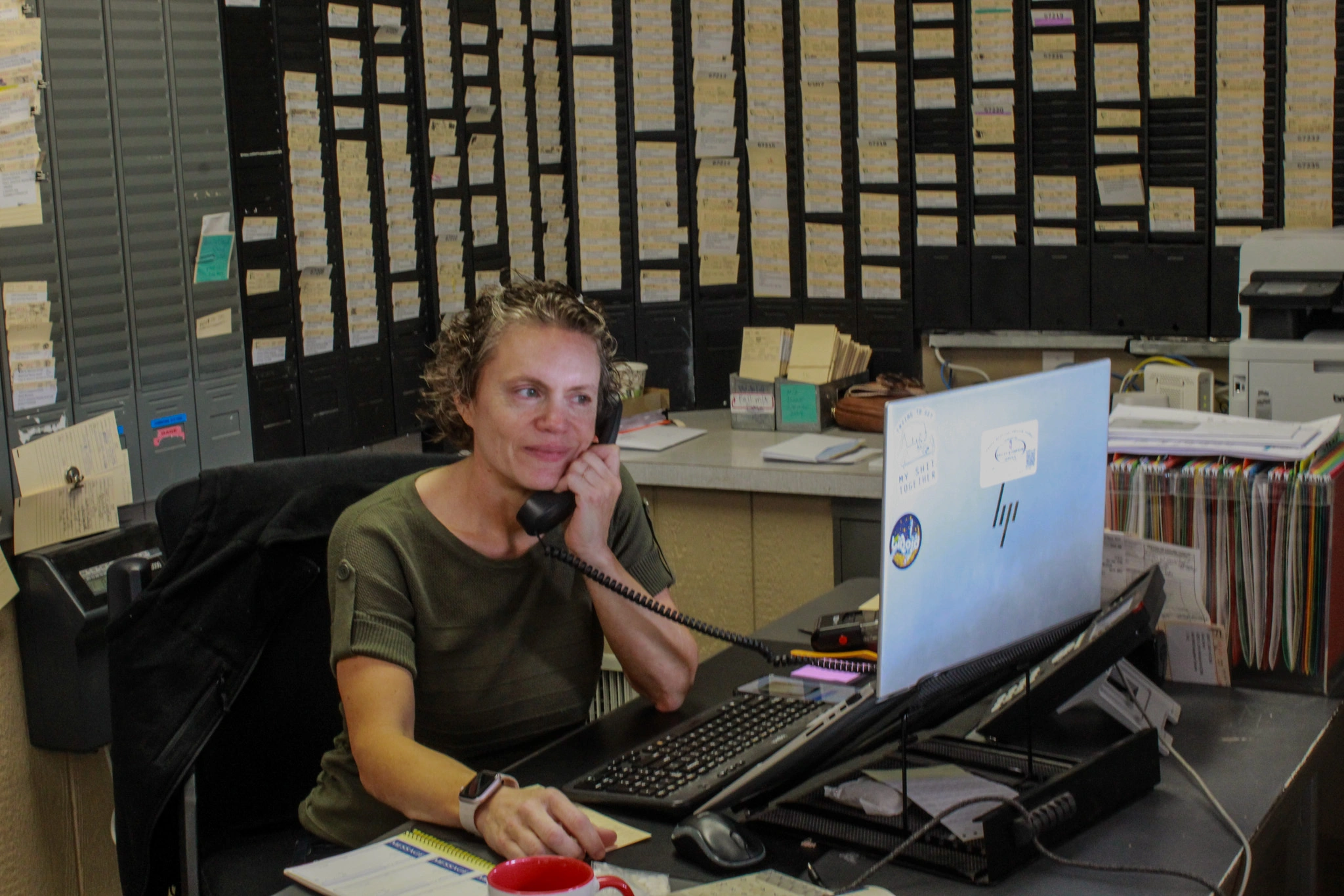 A woman with short hair sits in an office, talking on the phone while using a computer.