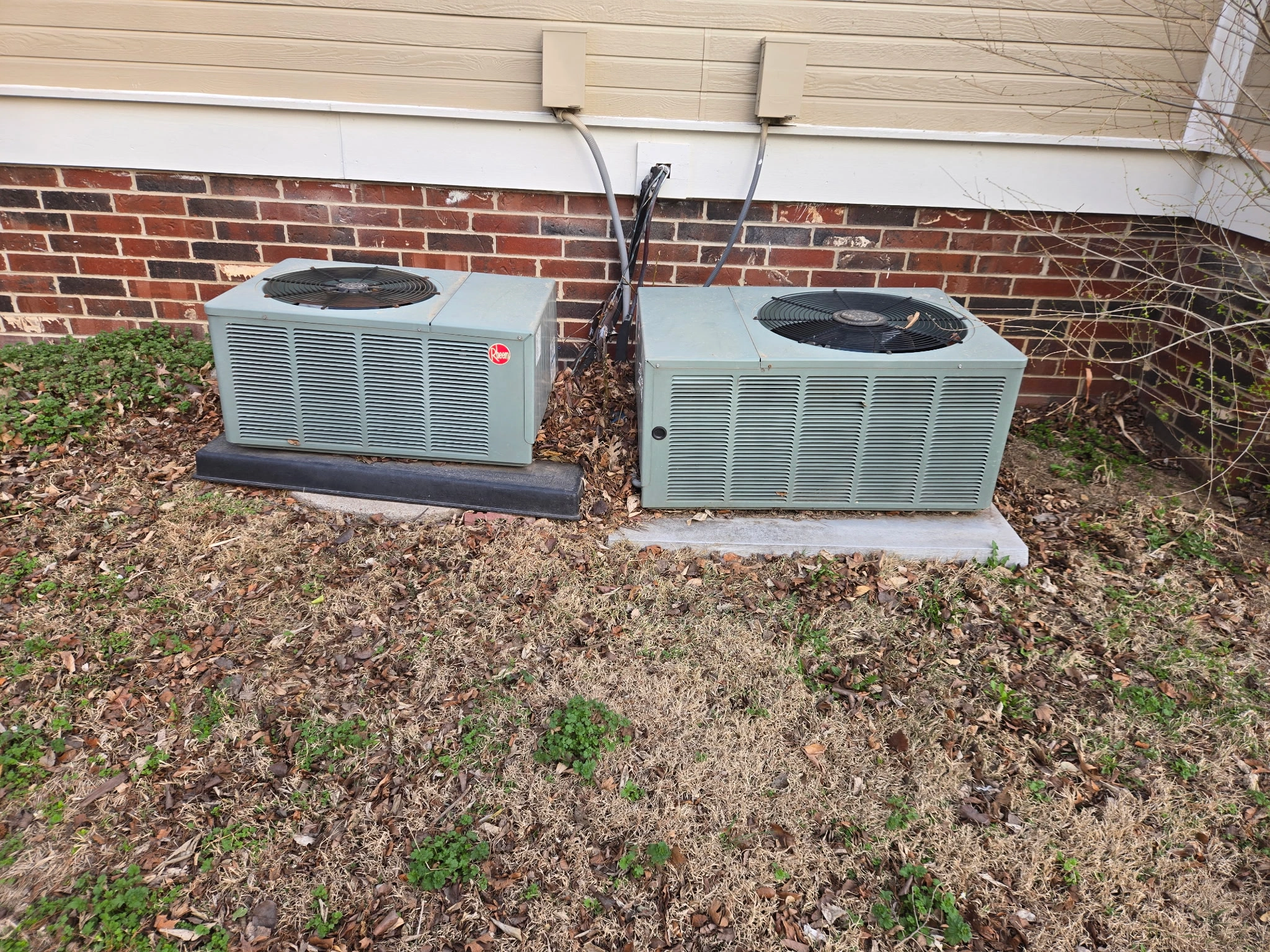 Two green outdoor air conditioning units on concrete slabs, set against a brick and siding wall