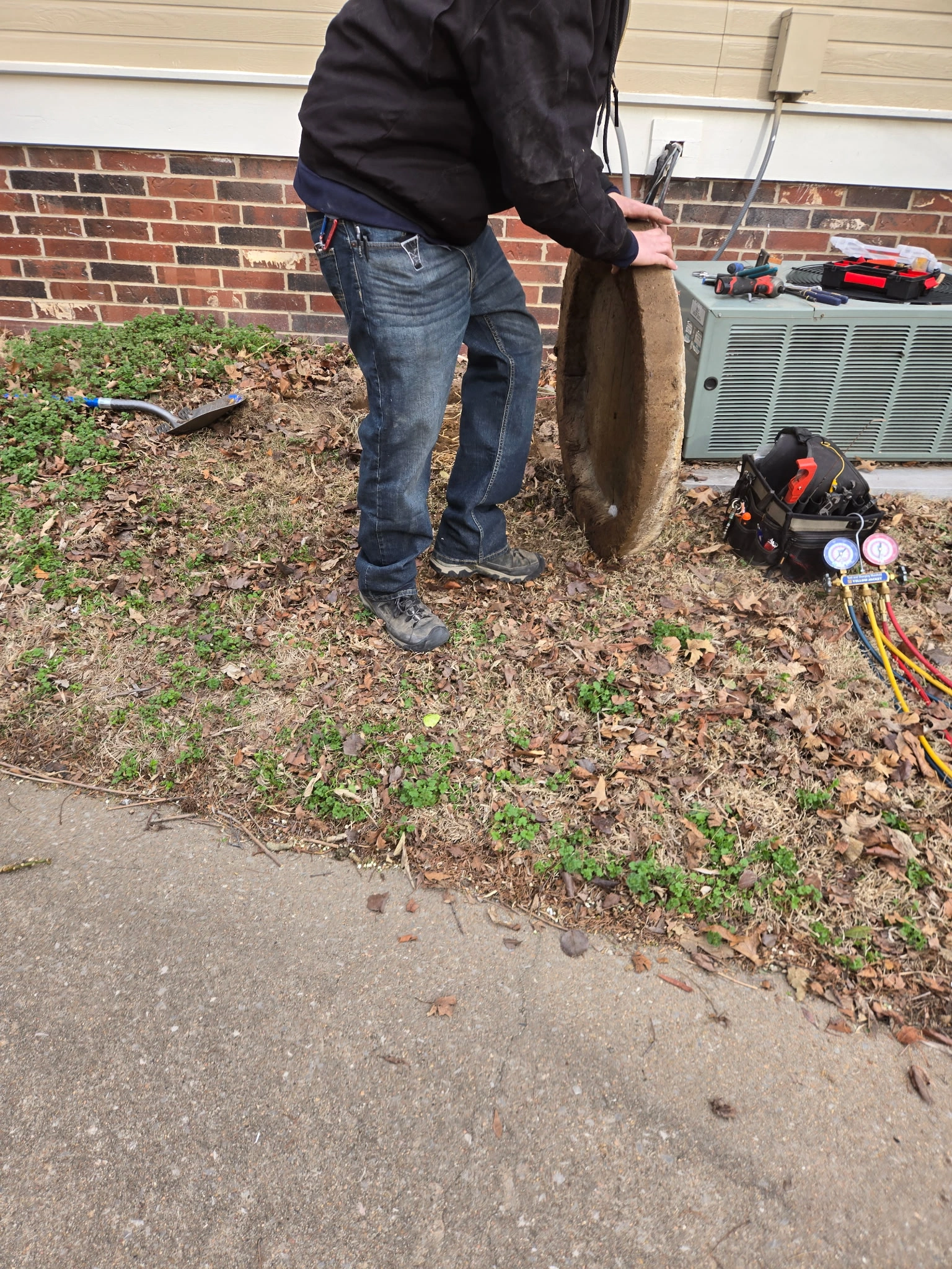 A person in jeans and a black jacket is lifting a circular concrete cover near an HVAC unit.