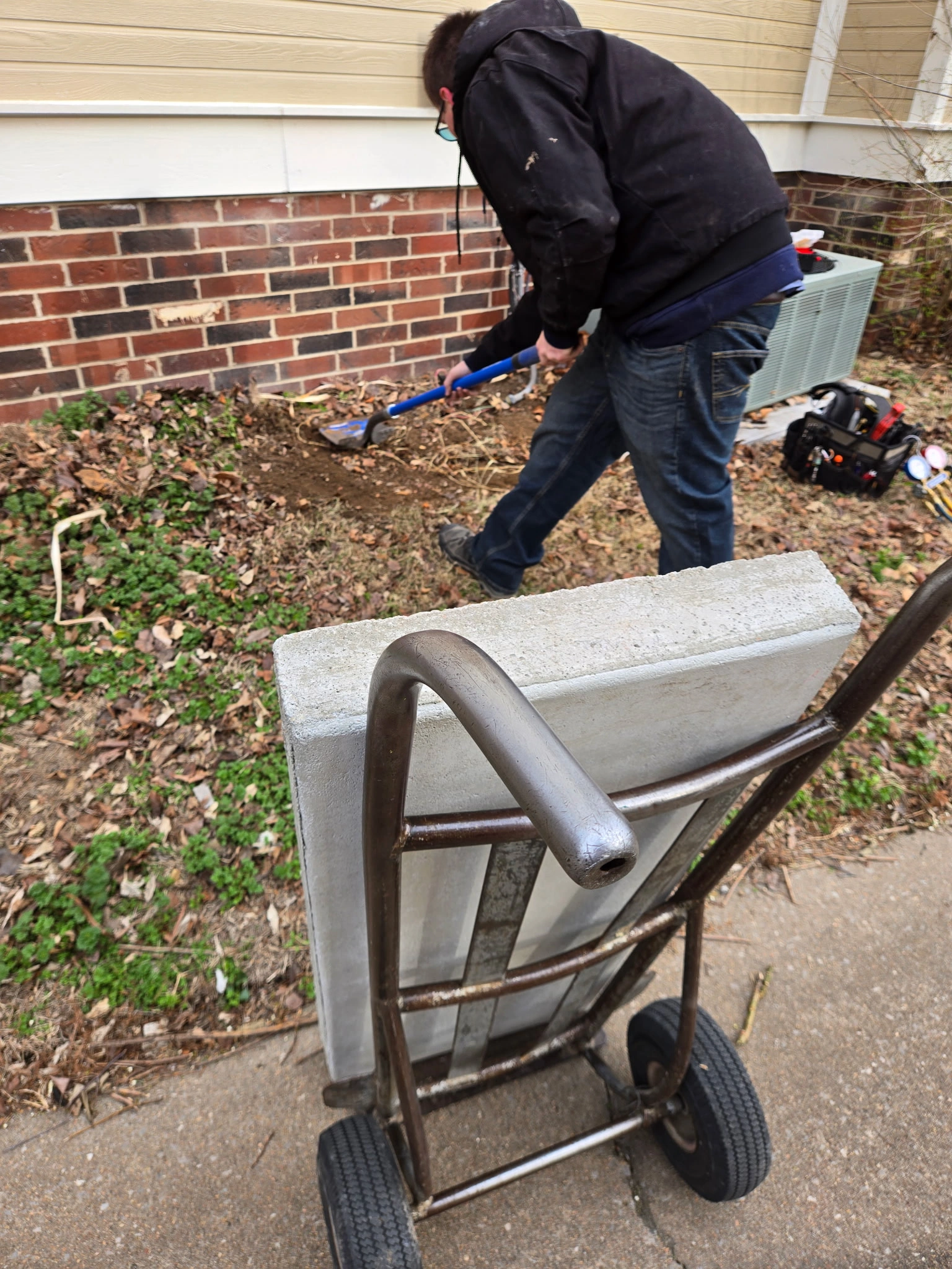 A person digs soil near a brick and siding wall with a shovel. In the foreground, a dolly carries a concrete slab.