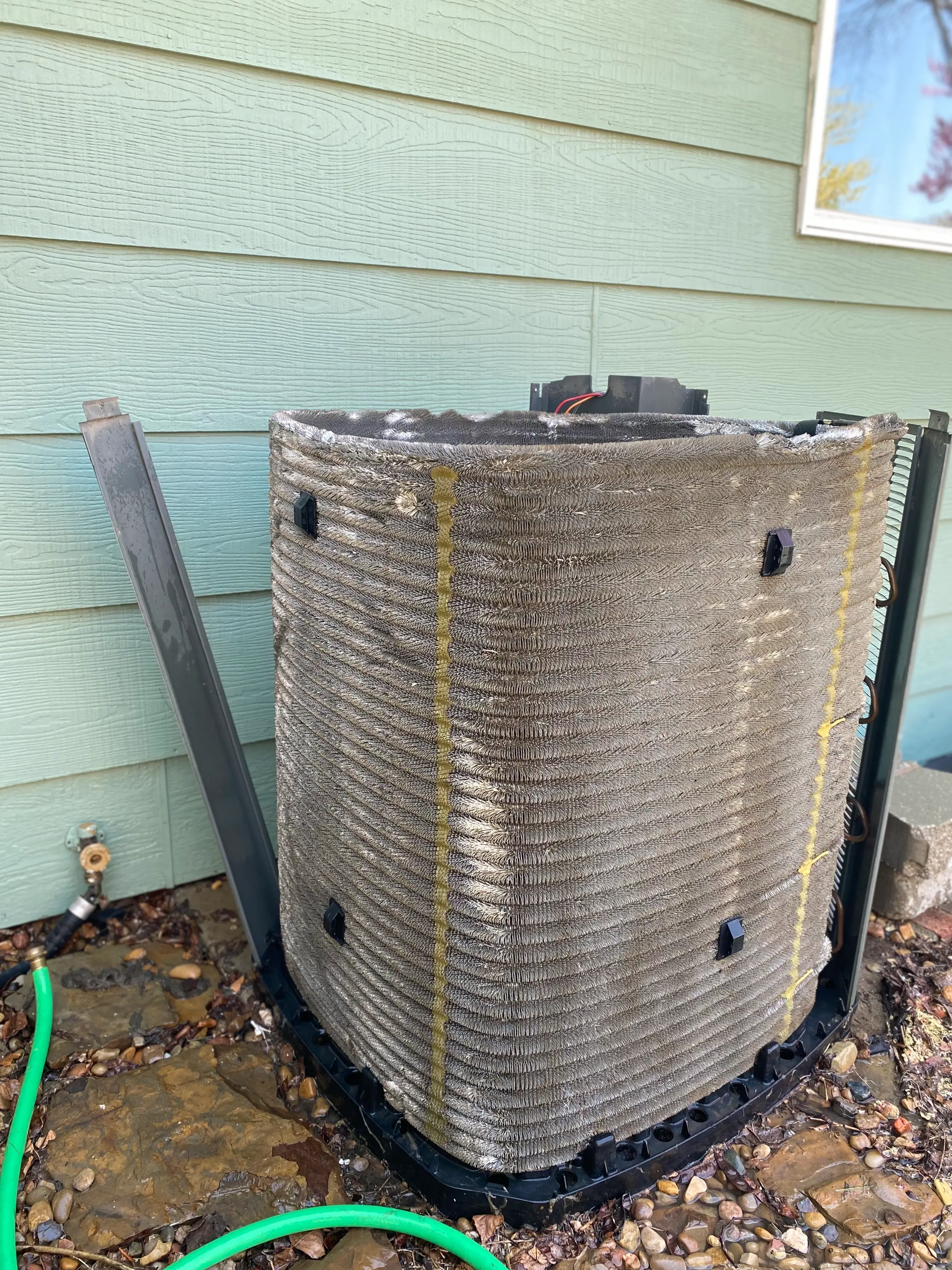 An outdoor air conditioning coil against a light green wall, partially disassembled, with a green hose on the rocky ground. 