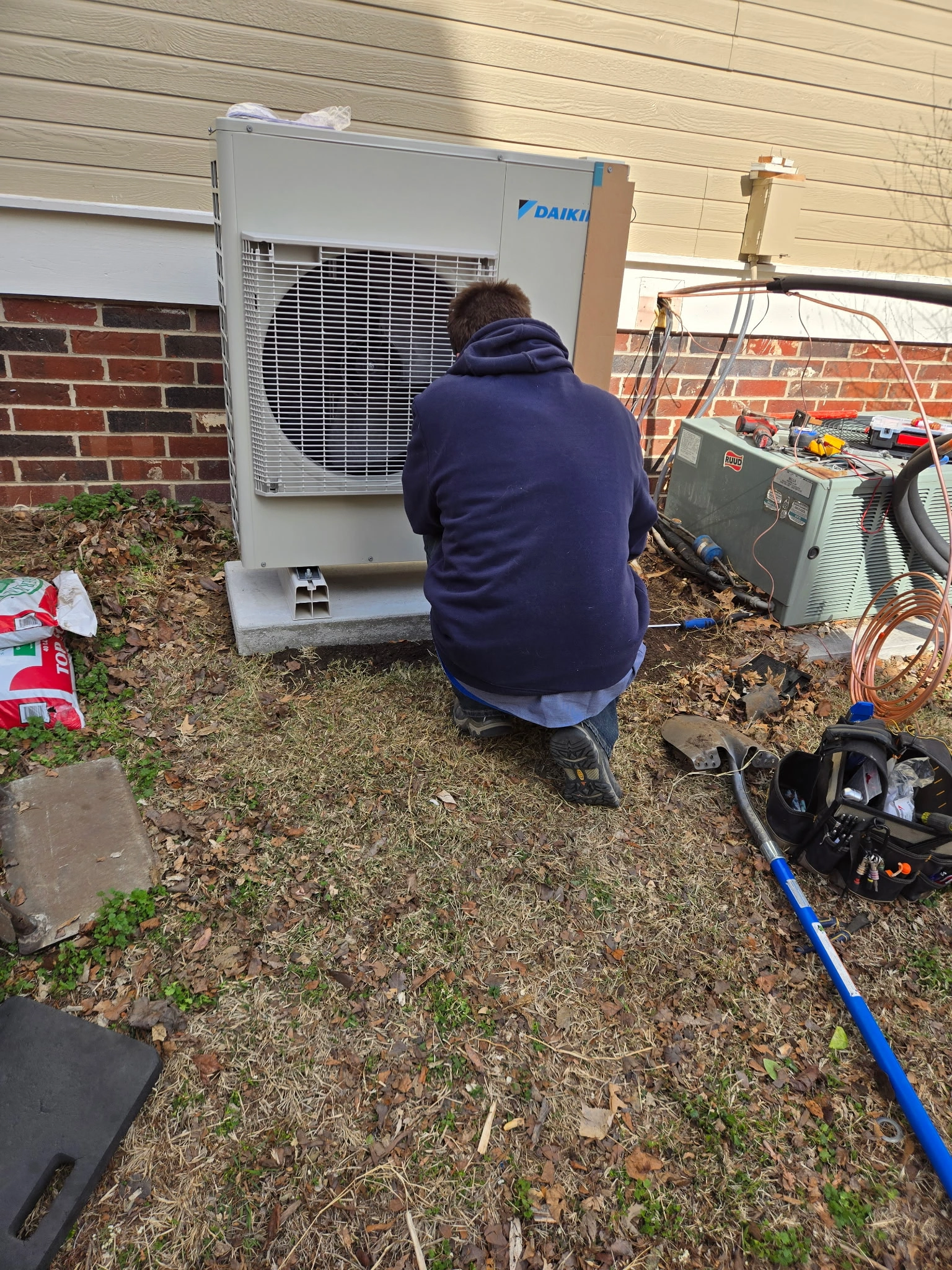 A person in a navy hoodie kneels on grass, working on an outdoor air conditioning unit next to a brick wall.