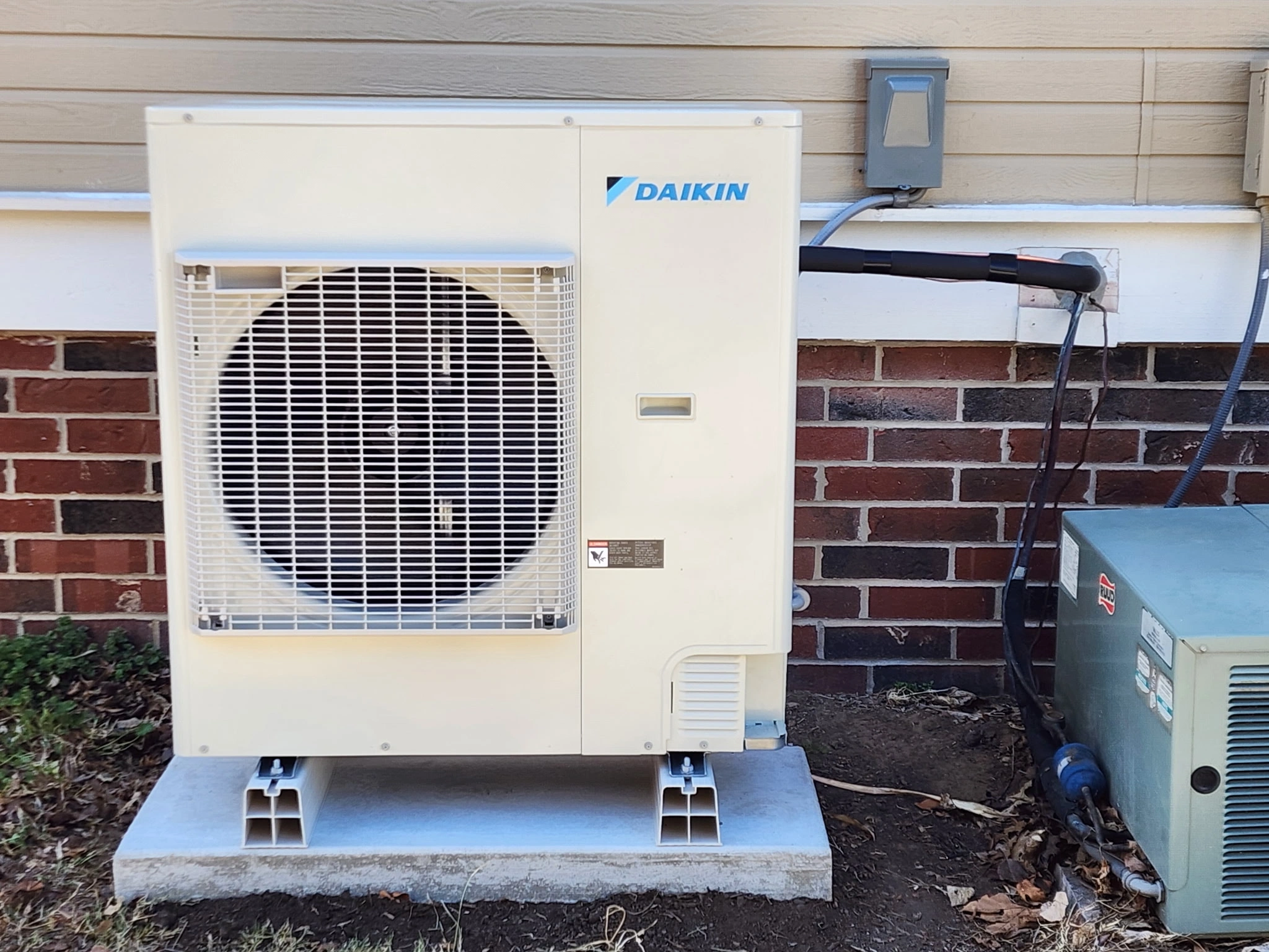 Outdoor air conditioning unit with a large front fan, branded Daikin, mounted on a concrete slab against a brick wall.