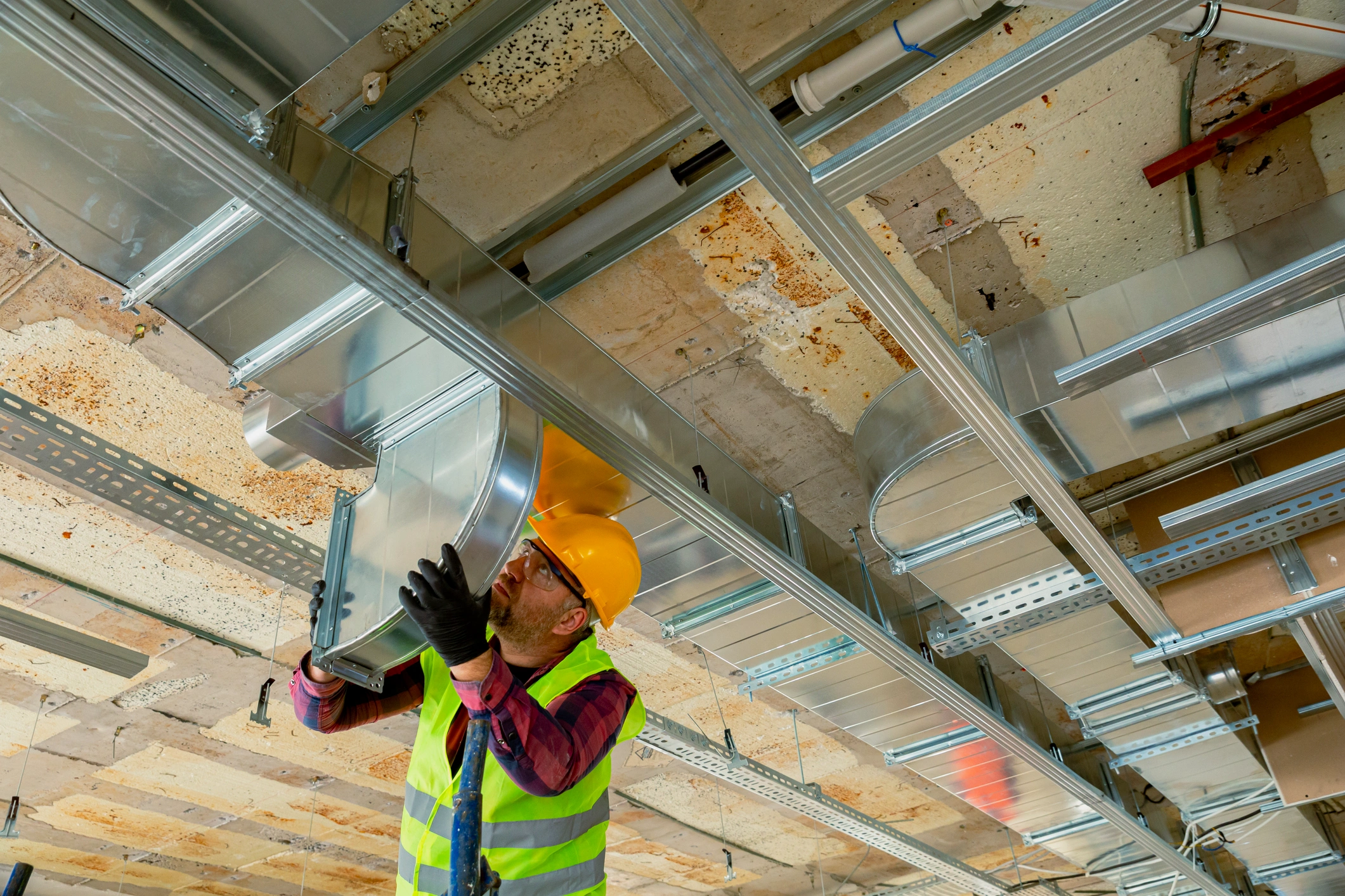 A construction worker in a hard hat and safety vest installs ductwork on a ceiling.