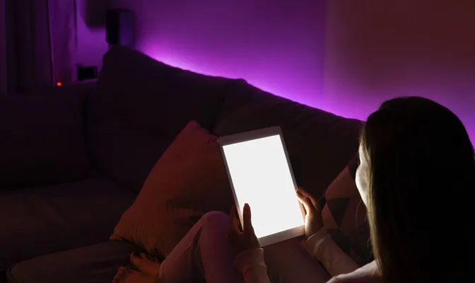 A woman is seen from behind, sitting on a couch in a dark room illuminated by purple mood lighting. They are holding a tablet with a bright, glowing white screen that lights up their face and the surrounding area.