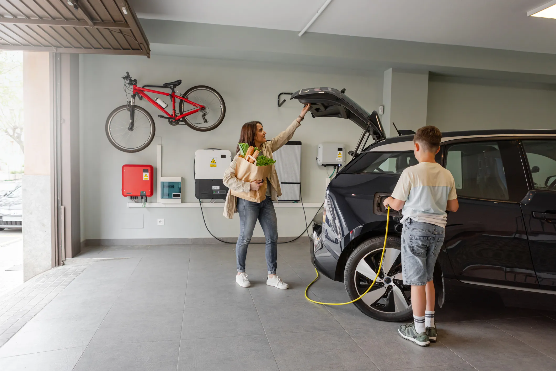 A woman holding a bag of groceries stands in an open garage, a bicycle is mounted on the wall behind her. She is reaching toward the back of a black electric vehicle (EV) that is plugged into a charging station on the wall. A young boy stands next to the c