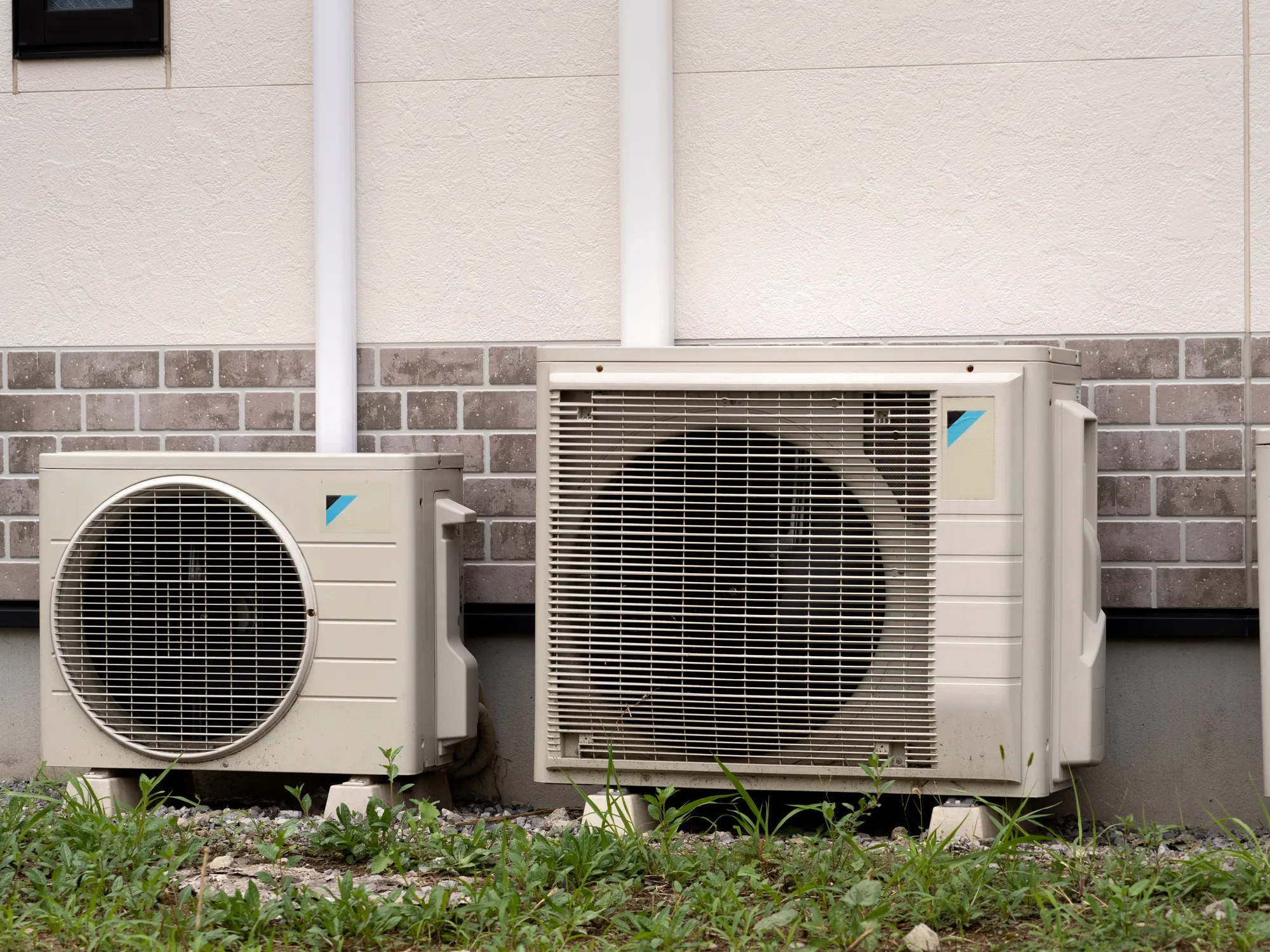 Two large, off-white exterior air conditioning or heat pump units sit on the ground next to a building with beige siding and brick-like accents. The units have visible fans behind metal grates and are connected to the building by white pipes.