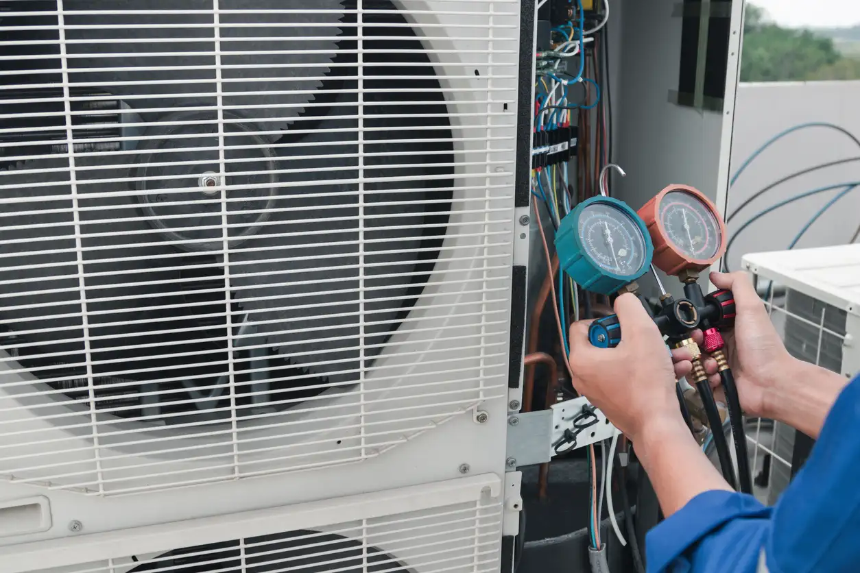 A professional HVAC technician is seen from the chest up, wearing a blue uniform, as they use a manifold gauge set to check the refrigerant pressure on a large outdoor HVAC unit. The two gauges, one with a blue dial and one with a red dial, are clearly visible. The unit's fan and the internal wires are exposed, showing that maintenance is underway. The image captures the technical nature of diagnosing a heat pump's performance.