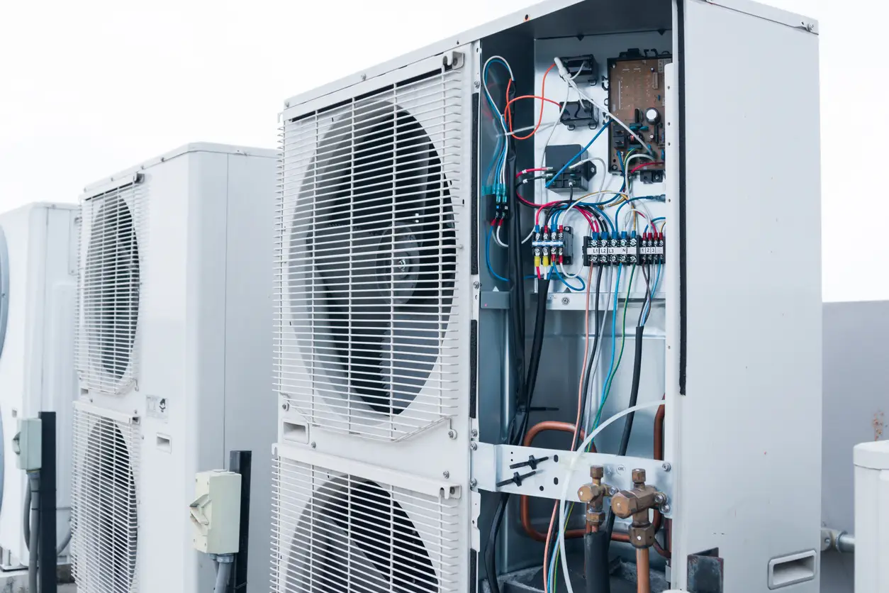 A professional heat pump technician is working on an outdoor HVAC unit, which has its side panel removed to expose a network of colorful wiring, circuit boards, and copper piping. The unit is one of several lined up on a rooftop. The image shows the complex internal components of a heat pump, highlighting the technical expertise required for maintenance and repair.