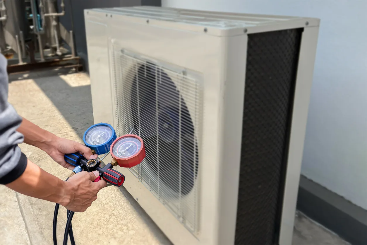 A pair of hands holds a manifold gauge set, with a blue dial and a red dial, connecting it to an outdoor heat pump unit. The heat pump is a rectangular, off-white unit with a fan visible behind a protective grid. The technician is performing a diagnostic check, likely to measure the refrigerant levels and pressure, on the HVAC system. The setting appears to be a rooftop or a concrete patio.