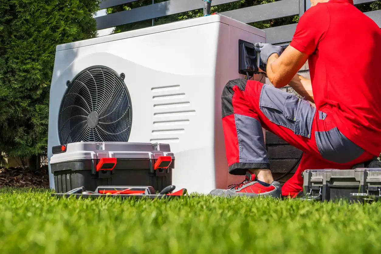 A ground-level shot of a professional HVAC technician, wearing a red shirt and gray pants, kneeling on a green lawn to service a large white heat pump unit. The technician is focused on the electrical panel of the unit. An open black and gray toolbox is positioned on the grass nearby, with various tools visible. The background includes a dark fence and green bushes, indicating an outdoor, residential setting.