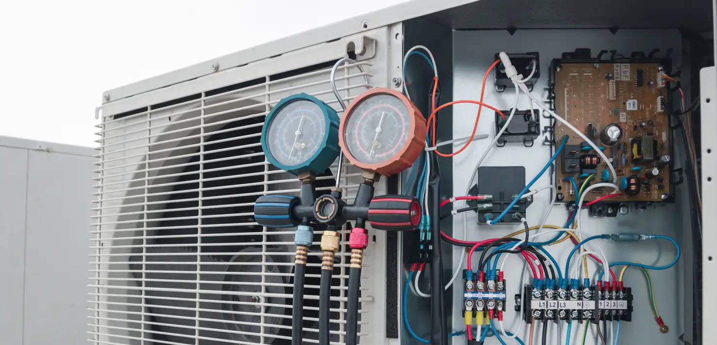 A close-up shot of an outdoor heat pump unit undergoing a professional diagnostic check. A manifold gauge set with two dials is attached to the unit's internal components. The side panel of the heat pump is open, revealing a complex arrangement of colorful wires, a circuit board, and other electrical parts. The heat pump's fan is partially visible behind a protective grille on the left. The image highlights the technical precision involved in HVAC maintenance.