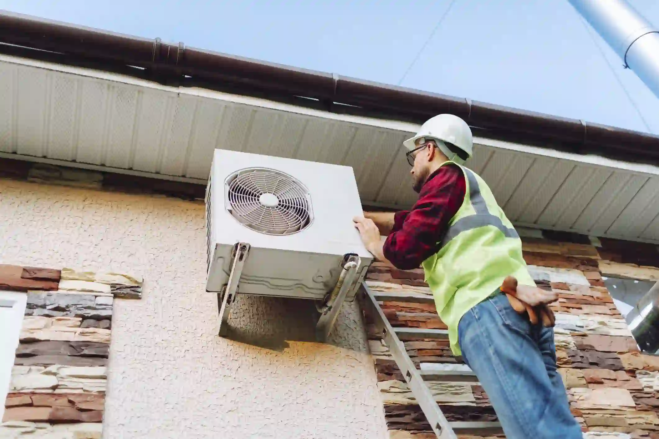 A professional technician, wearing a white hard hat, safety glasses, a red plaid shirt, and a high-visibility vest, is installing a white heat pump unit on the side of a building. He is standing on a ladder and is securing the unit to the wall. The building has a stone facade on the lower section and a light, textured finish above. A brown gutter runs along the roofline.