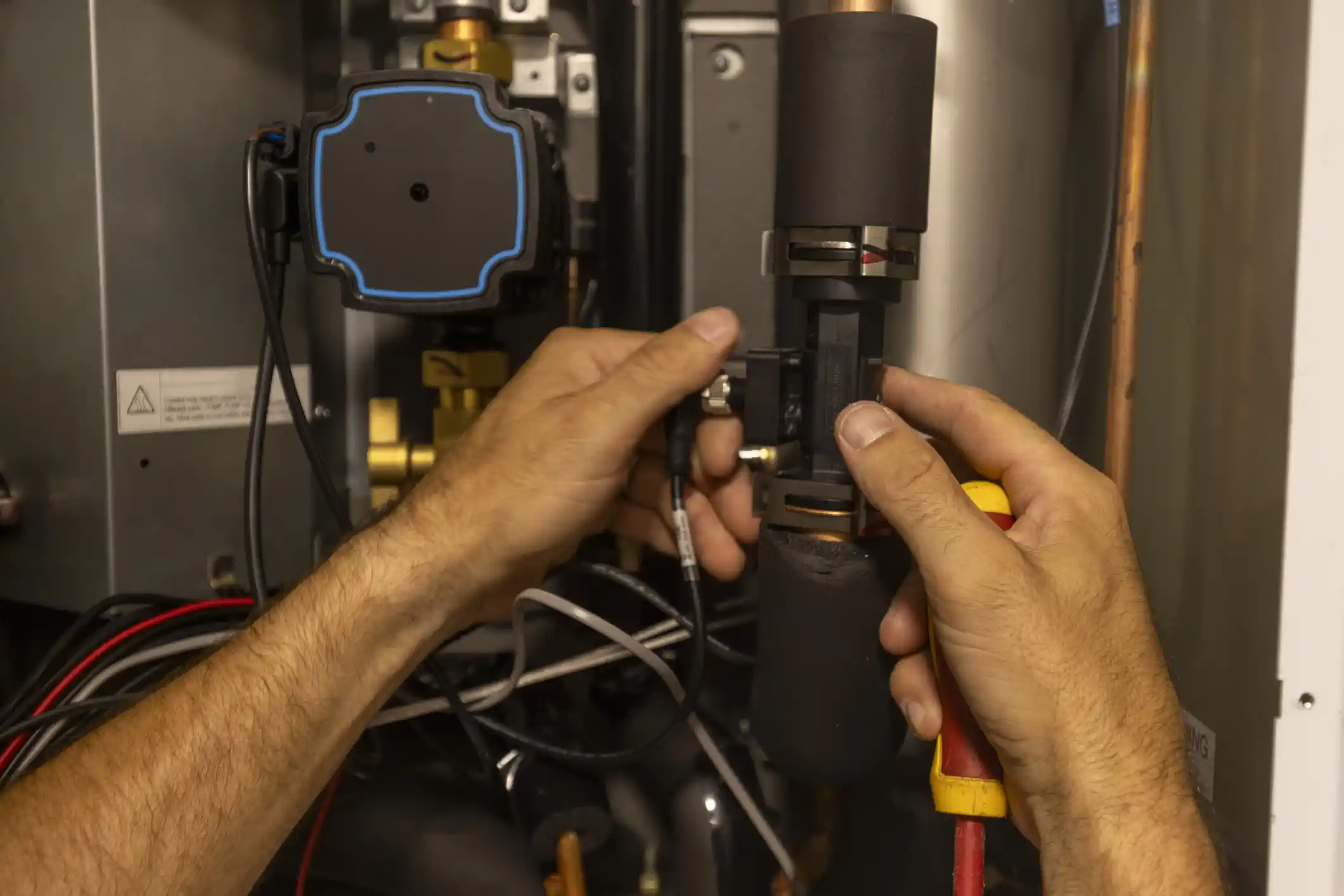 A close-up shot of a technician's hands, with a yellow and red tool, working on the internal components of a heat pump. The hands are connecting a black wire to a sensor or valve. Various pipes, insulated lines, and electrical wiring are visible within the unit's cabinet.