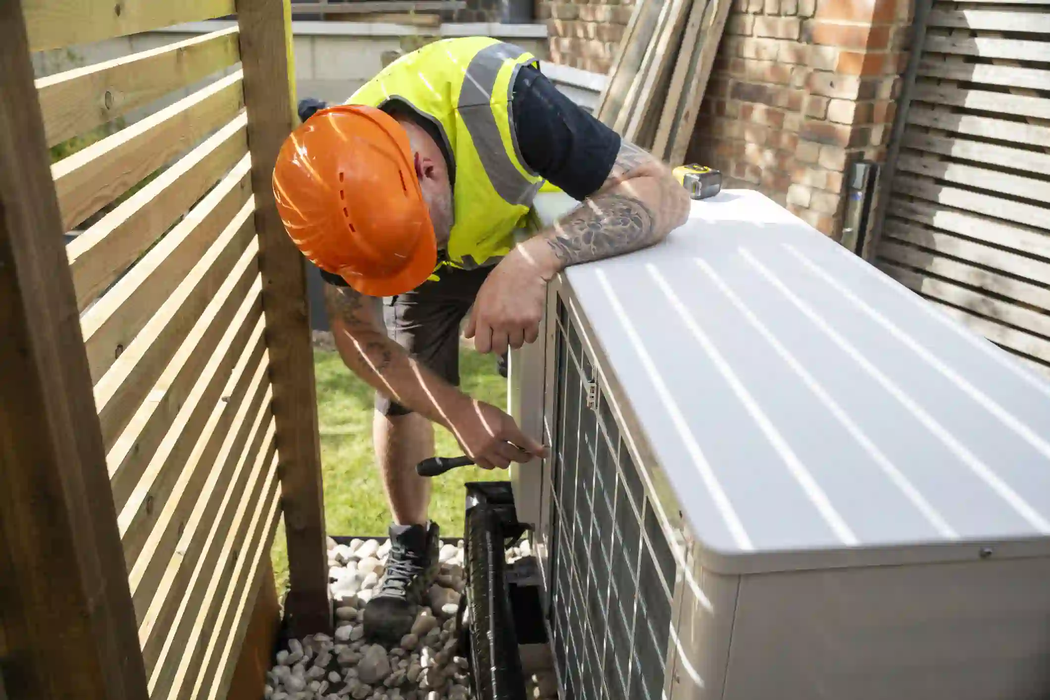 A professional technician with tattooed arms, wearing a black t-shirt, a high-visibility yellow vest, and an orange hard hat, is crouched down working on a white heat pump unit. The unit is situated on a bed of white stones next to a wooden fence. The technician is using a screwdriver to work on a panel on the side of the unit. A brick wall is visible in the background.
