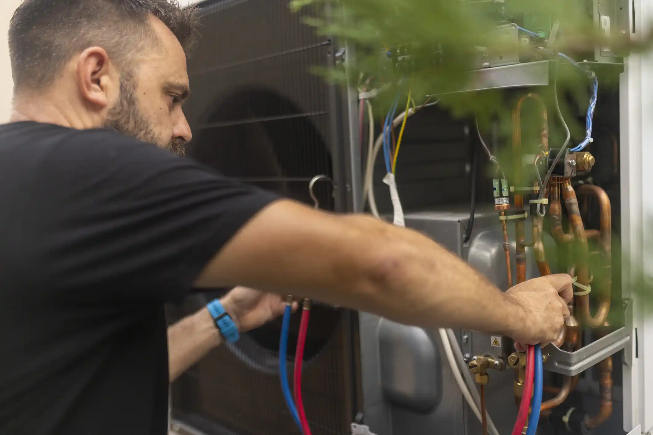 A close-up side view of a male HVAC technician wearing a black t-shirt. He's working on the internal components of an outdoor heat pump unit, holding a red and a blue hose connected to the system. His other hand is on the copper pipes and valves. The background is slightly out of focus, with some greenery in the foreground.
