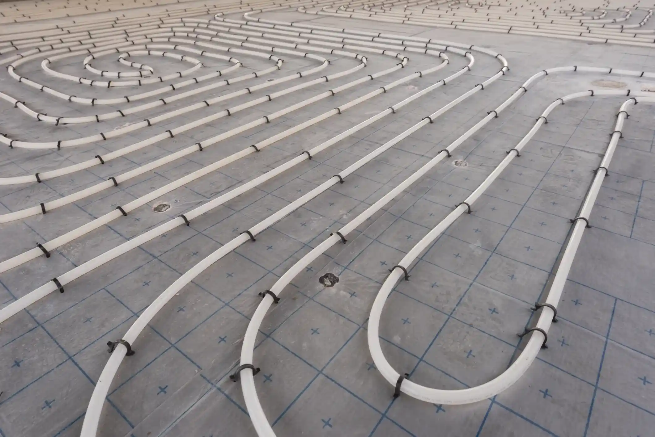 A wide-angle view of a construction floor with a looping pattern of white radiant heating pipes secured on a gray, gridded mat.