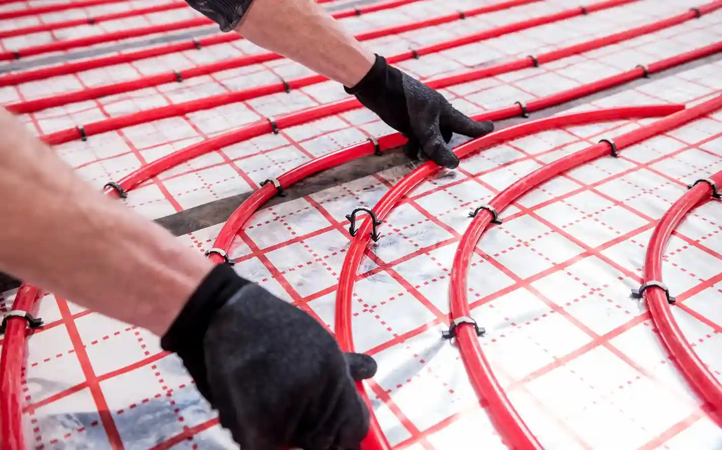 A close-up of a worker's gloved hands installing red radiant heating pipes on a reflective, gridded matting on a floor.