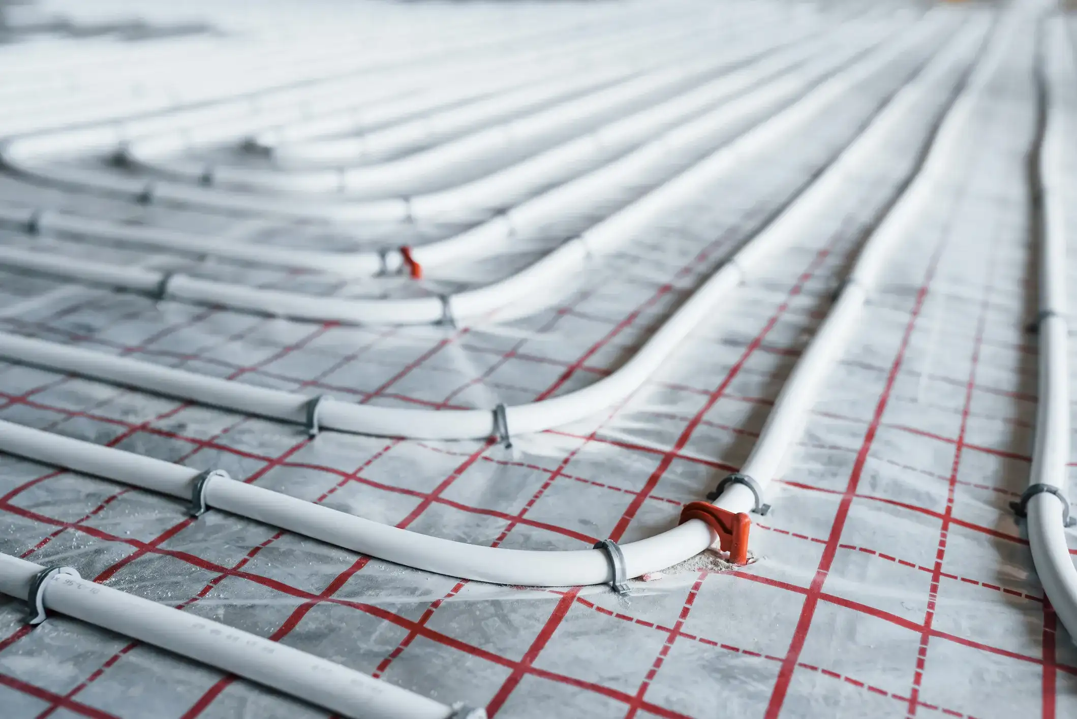 A close-up, low-angle shot of white radiant heating pipes laid on a red gridded mat, with some pipes secured by bright red clips.