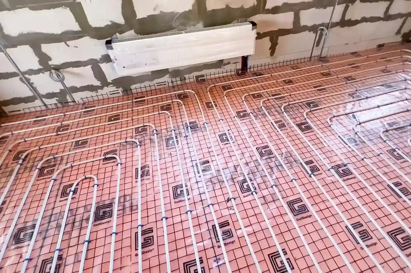 A wide shot of a room under construction, showing a network of white radiant heating pipes laid on a pink insulation mat on the floor.