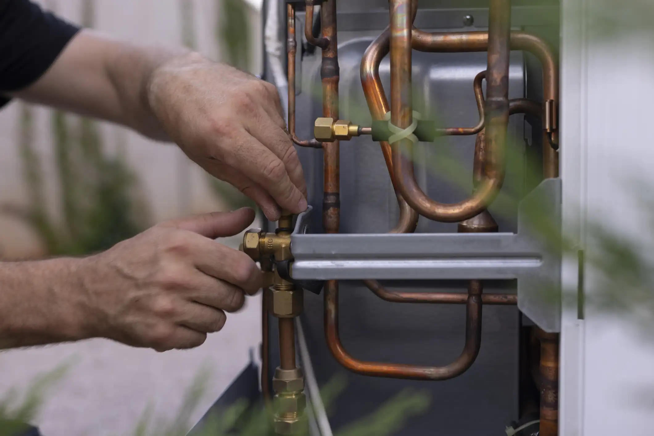 A close-up shot shows a professional technician's hands adjusting a brass fitting on the copper piping inside a heat pump unit. The technician is wearing a black long-sleeved shirt. The complex network of pipes and fittings is clearly visible, with some components wrapped in a white covering. The background is a blurry green.