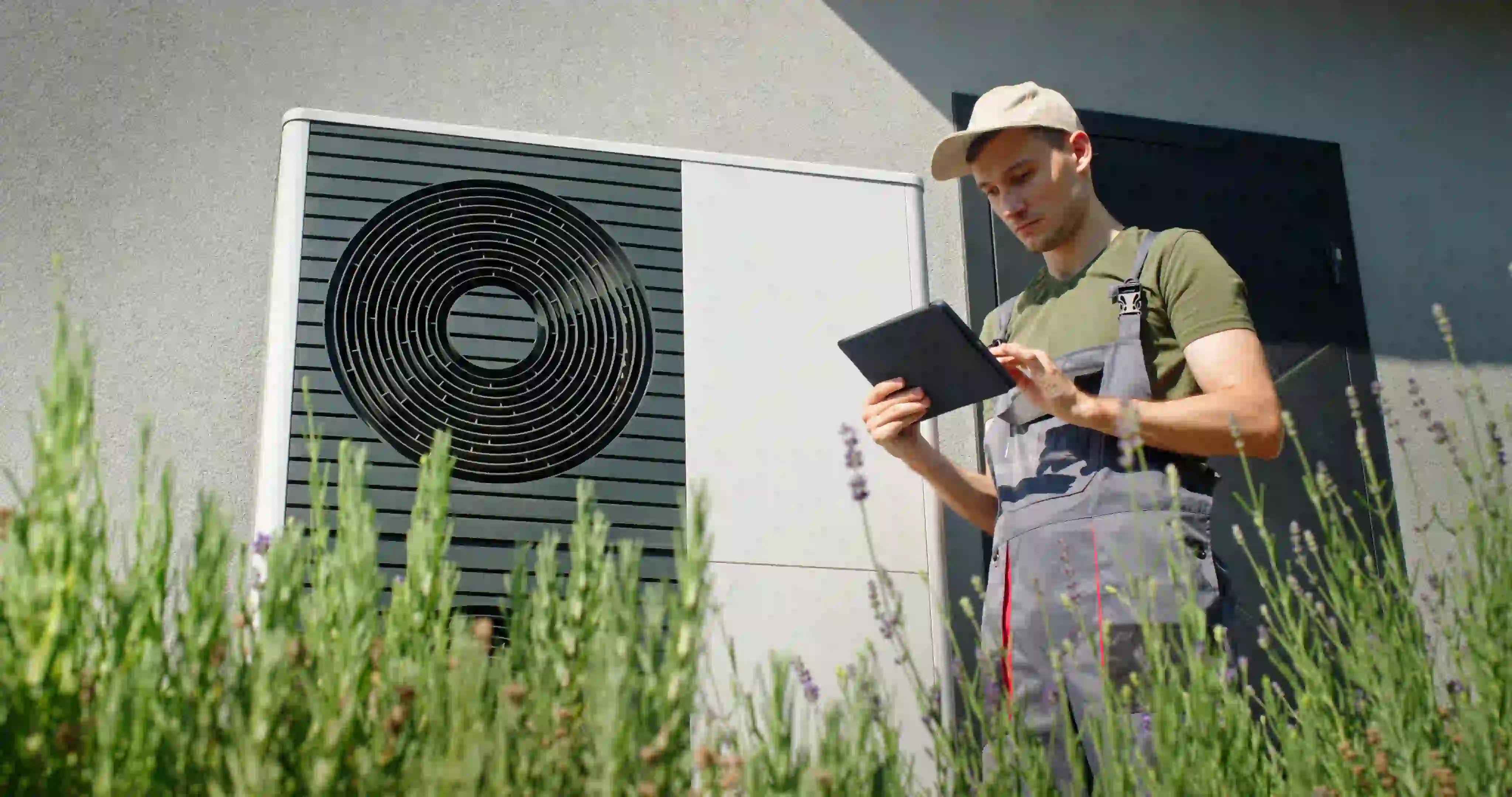 A professional technician, dressed in a grey uniform and a beige hat, stands outside a modern building. He is looking at a tablet while inspecting a large, white heat pump unit mounted on the wall. The foreground shows a patch of lavender.